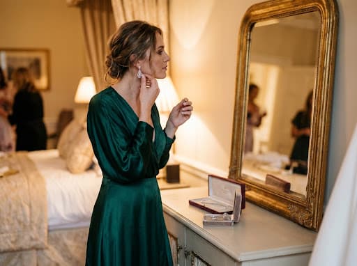 A woman in a green silk dress fastening a drop earring at a gilded mirror